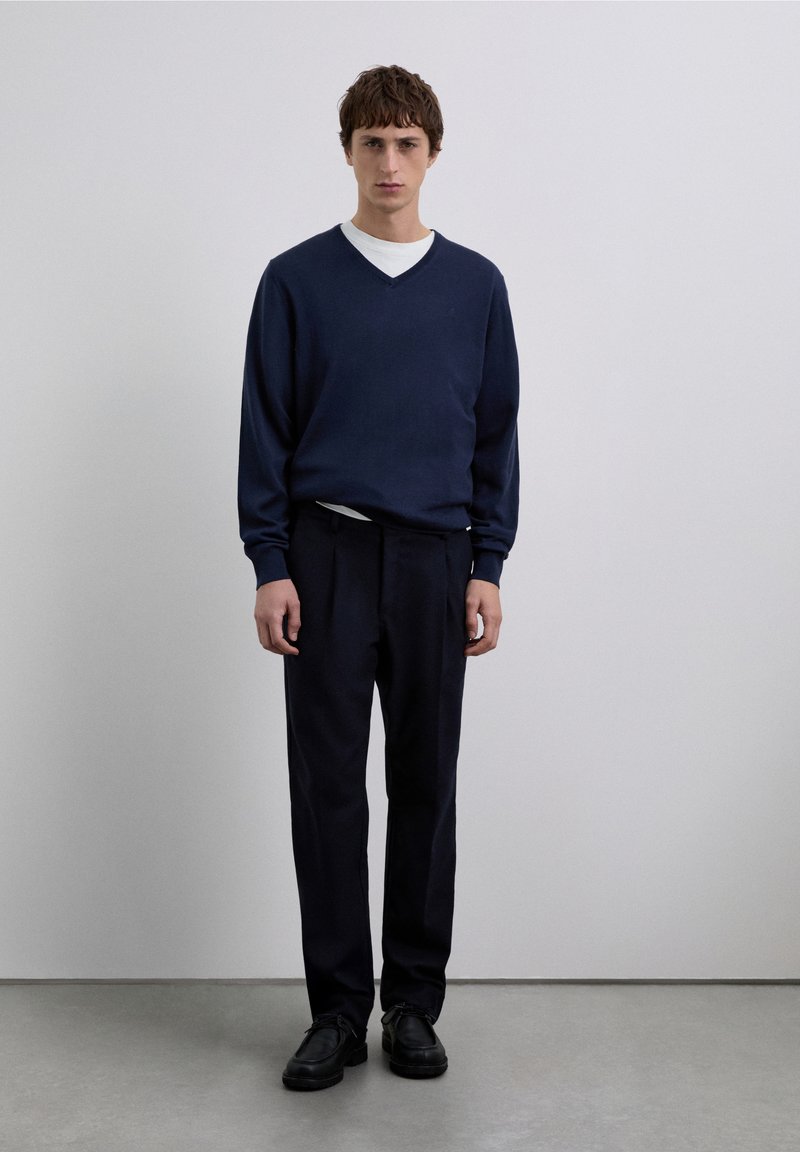 Marineblauwe gebreide trui met een V-hals, gedragen over een wit overhemd, gecombineerd met donkere broek en zwarte schoenen, staand tegen een effen achtergrond.