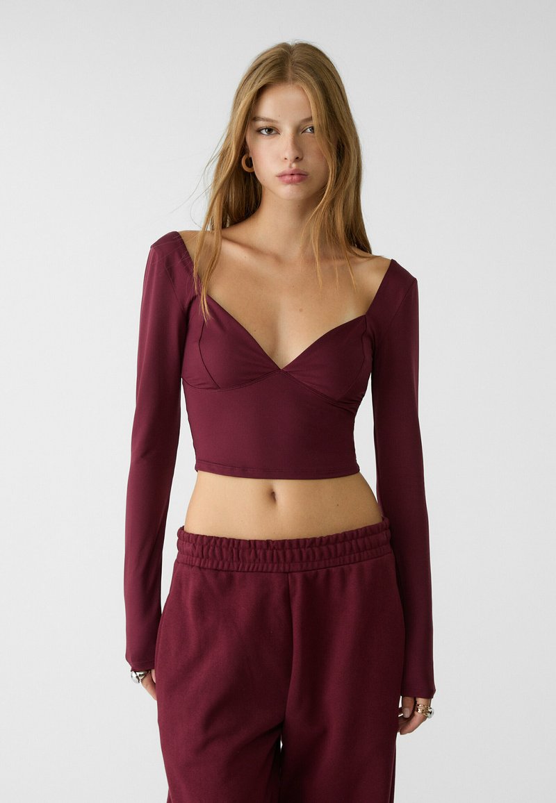 Crop top a maniche lunghe color borgogna con scollatura a V profonda, design aderente e texture liscia, abbinato a pantaloni tuta coordinati con vita elastica.