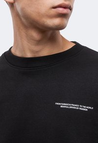 Κοντινό ενός ατόμου που φοράει μαύρο πουκάμισο με λευκό κείμενο στο στήθος που λέει "FROM FARMÁCIA FRANCO TO THE WORLD BENFICA, DRIVEN BY PASSION".