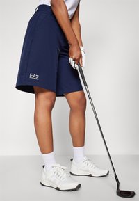 Mornarske golf hlače s logotipom, s prilagojenim krojem, ravnim prednjim delom in žepi. Uporabljene z belo srajco in golf čevlji. Golf palica v roki.