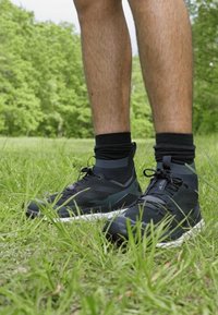Schwarze Sportschuhe mit einem hohen Schaft, Obermaterial aus strukturiertem Stoff, mit grauen Akzenten und einer weißen Sohle. Getragen mit schwarzen Socken im Knöchel auf Gras.