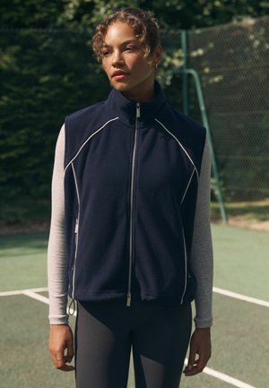 Laevast sinine fliisvest kõrge kraega, hõbedase lukuga ja valge tikandiga, kantud helerohelise pikkade varrukatega särke peal, koos tumedate retuusidega.