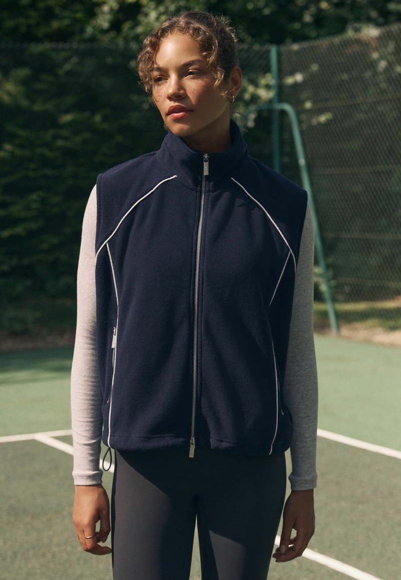 Laevast sinine fliisvest kõrge kraega, hõbedase lukuga ja valge tikandiga, kantud helerohelise pikkade varrukatega särke peal, koos tumedate retuusidega.