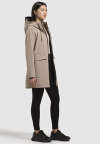 Beige hættejakke med lynlås foran, side lommer og justerbare snore. Båret over sorte leggings og sorte træningssko.