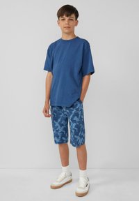 Blauwe katoen T-shirt met korte mouwen, gecombineerd met knieboven blauwe camouflageshorts en witte sneakers met tan accenten en witte sokken.