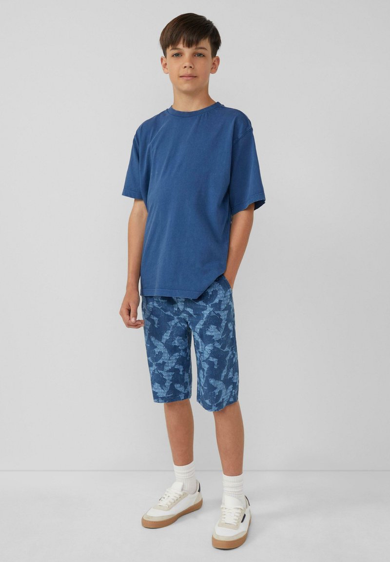 Blauwe katoen T-shirt met korte mouwen, gecombineerd met knieboven blauwe camouflageshorts en witte sneakers met tan accenten en witte sokken.