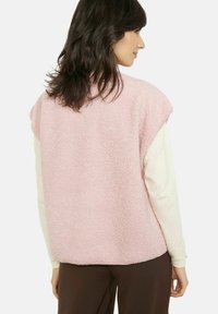 Rozā flīsa veste ar teksturētu virsmu, īsām cepures piedurknēm un apaļu maliņu. Valkāta virs krēmkrāsas garroku topa, attēlota no aizmugures.