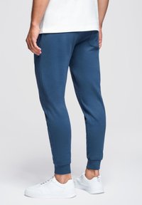 Blauwe joggerbroeken met ribgebreide manchetten, zijzakken en een zachte textuur, gecombineerd met witte sneakers op een neutrale achtergrond.