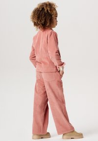 Rosa Cordjacke und Hose im entspannten Schnitt, mit strukturierten Streifen, aufgesetzten Taschen und beige Slippern.
