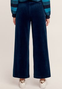 Brede navy corduroy broek met een gestructureerd oppervlak. Bevat gestikte zakken en een hoge taille. Gecombineerd met witte sneakers met gouden accenten.