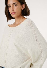 Invito a beige chiaro in versione oversize, realizzato in morbido tessuto a maglia. Presenta un ampio scollo a barchetta, spalle a caduta e dettagli ad occhio decorativi lungo le maniche.