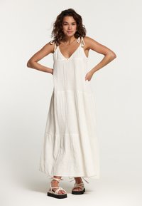 Robe maxi blanche sans manches avec un décolleté en V profond, des bretelles à nouer, une texture froncée et un design à volants. Associée à des sandales épaisses.