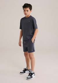 T-shirt à manches courtes gris foncé et shorts assortis, fabriqués en tissu doux. Présente un petit logo sur le t-shirt et des poches dans les shorts. Porté avec des baskets noires et grises.