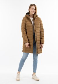 Bruine gewatteerde lange jas met een ritssluiting en capuchon. Zachte textuur, gequilt ontwerp, aangevuld met lichtblauwe spijkerbroek en beige sneakers.