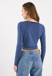Langermet, cropped topp i marineblå med blonder langs kanten og halsen, brukt sammen med høyt liv lyseblå denimjeans.