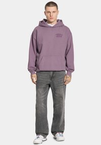 Fioletowa bluza z kapturem i przednią kieszenią, zestawiona z wyblakłymi czarnymi dżinsami. Dopełnieniem stroju są białe trampki z niebieskimi akcentami.