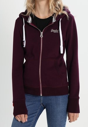 Mujer con sudadera burdeos con cremallera y cordones blancos y vaqueros azules, de pie frente a un fondo claro y liso.