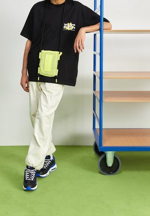 Camiseta negra de manga corta con gráfico, bolso bandolera verde lima, pantalones cr kremados y zapatillas negras con acentos azules están sobre un suelo verde.