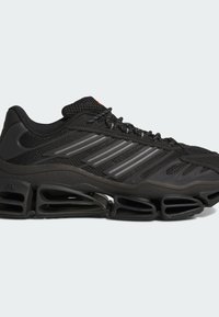 Baskets de sport noires avec tige en mesh, bandes réfléchissantes, semelle épaisse segmentée et lacets visibles sur un fond clair uni.