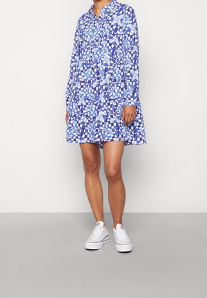 Kvinde iført en langærmet, blå og hvid kjole med geometrisk mønster, parret med hvide platform-sneakers, stående foran en ensfarvet baggrund.