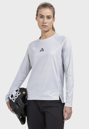 Frau in weißem Langarmshirt und schwarzer Hose hält einen schwarzen POC-Helm und steht vor einem neutralen hellen Hintergrund.