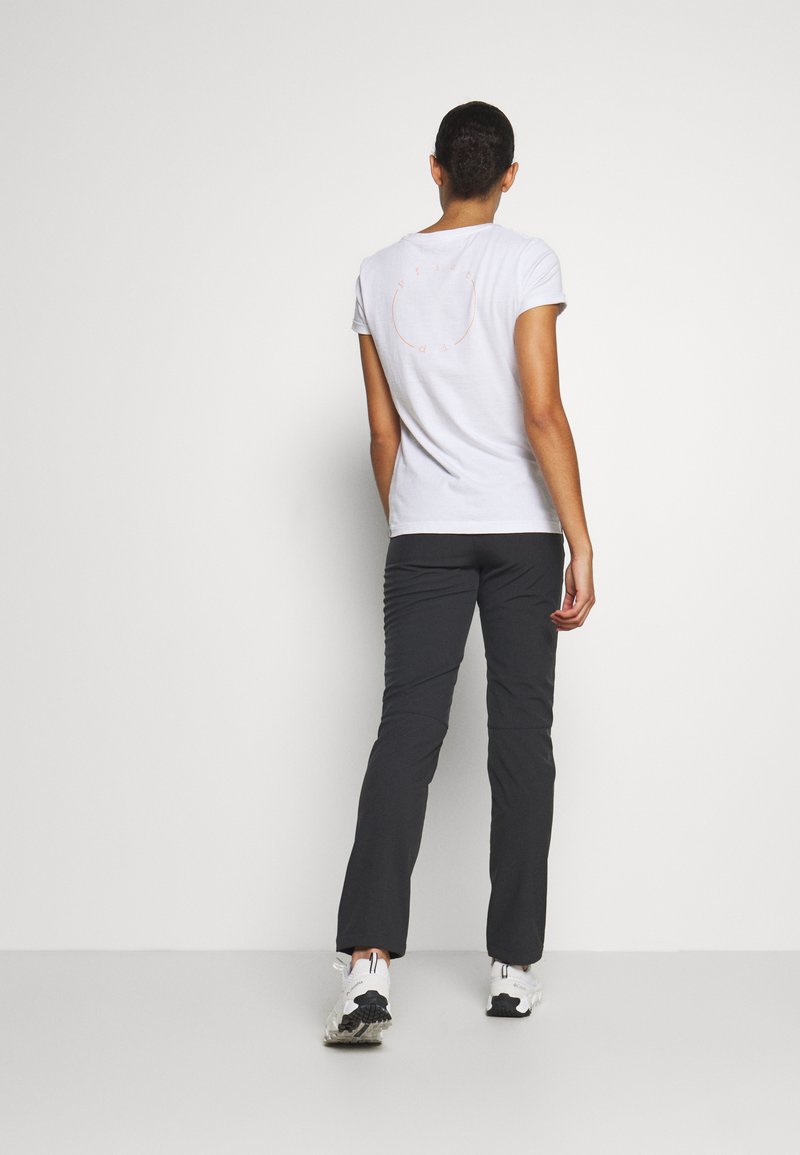 Witte t-shirt met korte mouwen en een ronde oranje afbeelding op de achterkant, gecombineerd met zwarte broeken en witte sportschoenen.