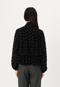 Black polka dot blouse with layered ruffle detail, made of sheer fabric, featuring long sleeves and a high collar. Textured finish.