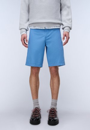 Mężczyzna w jasnoniebieskich szortach do kolan, szarych skarpetach, czarnych butach z czerwonymi sznurowadłami oraz jasnoszarym swetrze nałożonym na pasiastą koszulę.