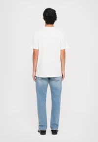 Mann med krøllete mørkt hår står med ryggen til iført en enkel hvit t-skjorte, lyseblå jeans og svarte sko mot en hvit bakgrunn.