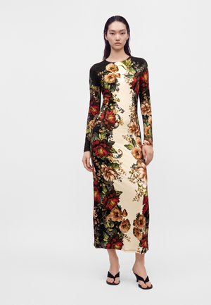 Donna con un lungo vestito floreale con pannelli neri e beige, in piedi a piedi nudi su sfondo bianco semplice.