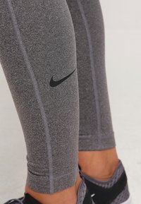 Jambes vêtues de leggings gris Nike avec un logo noir sur le mollet et de chaussures de sport noires avec des accents violets sur un sol clair.
