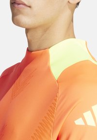 Oranje sportshirt met een hoge kraag, voorzien van gele accenten en textuurpatronen op de stof, ontworpen voor prestaties.