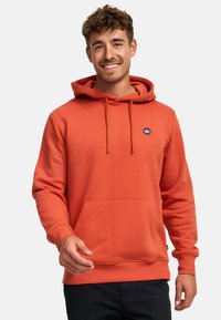 Felpa arancione realizzata in materiale morbido, dotata di tasca a marsupio, cappuccio regolabile con cordino e un patch rotondo con logo sul petto.
