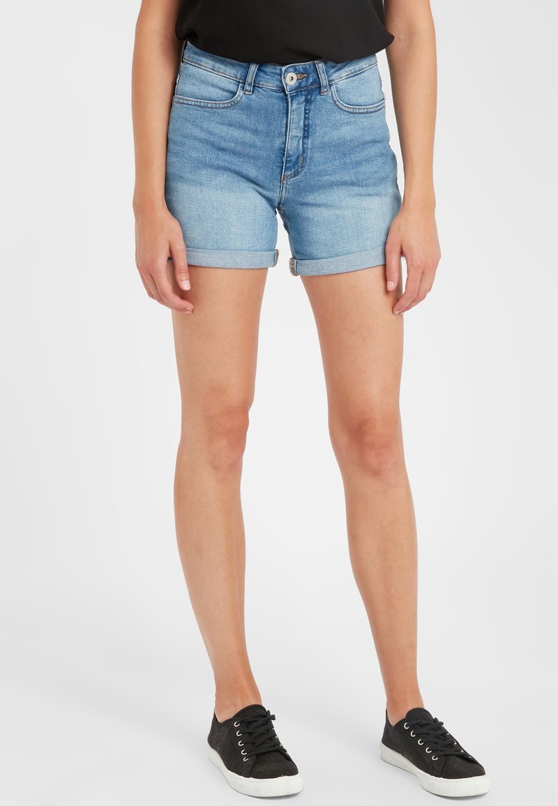 Femme portant un short en jean bleu clair roulotté, un haut noir et des baskets noires à semelles blanches, debout devant un fond uni.