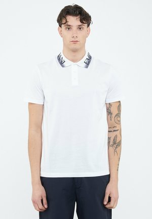 Giovane uomo con capelli corti e ricci che indossa una polo bianca con scritta nera del logo sul colletto e pantaloni scuri, mostrando tatuaggi dettagliati sulle braccia.