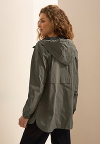 Chaqueta ligera de color verde oliva con capucha, cordón y ventilación trasera; presenta una textura suave y un dobladillo redondeado.