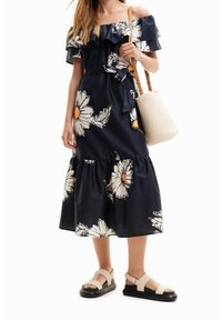 Laivansininen off-the-shoulder-mekko, jossa on valkoinen kukkakuosi, röyhelökaula-aukko, sidottu vyötärö ja levenevä helma. Kankainen beige laukku ja sandaalit täydentävät asun.