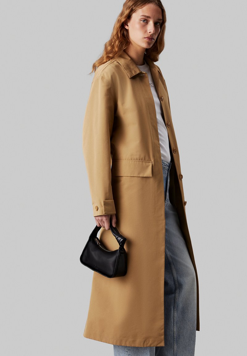 Kamelový trench coat s límcem, předními knoflíky a bočními kapsami. Modelka drží malou černou kabelku a nosí světle modré džíny.