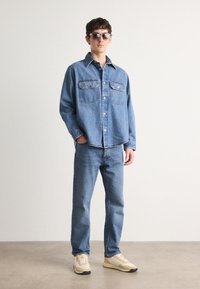 Denim shirt with two front pockets, light blue color, paired with light blue jeans. Wears sunglasses and beige sneakers with dark soles.