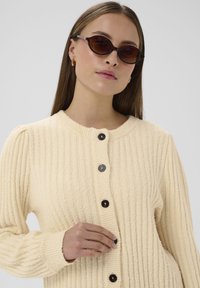 Krēmkrāsas ribotais adījuma kardigāns ar piecām tumšām pogām, apaļu izgriezumu un piegulošām piedurknēm. Modelis valkā vīna krāsas kaķa acs saulesbrilles.