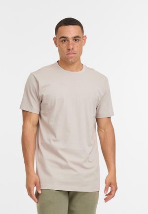 Un jeune homme avec des cheveux courts et des taches de rousseur portant un t-shirt beige à manches courtes et un pantalon vert olive devant un fond blanc uni.