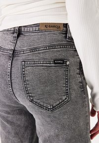 Vaqueros de mezclilla gris con una textura suave, que presentan un bolsillo trasero con una etiqueta negra y una etiqueta de cuero marrón marcada con "GARCIA" en la pretina.