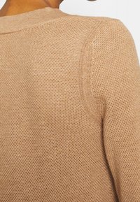 Gros plan sur un pull en maille texturée de couleur beige, mettant en évidence l'épaule et la partie supérieure du dos, porté par une personne aux cheveux courts.