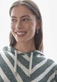 Grå och krämrandig hoodie med dragsko, tillverkad av mjukt material. Modellen bär silverörhängen med ringar och ler mot en ljus bakgrund.