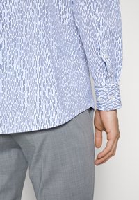 Camisa de manga larga con estampado azul y blanco, con líneas verticales, puños con botones y dobladillo redondeado; combinada con pantalones gris claro.