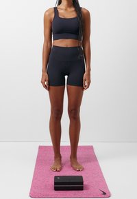 Kobieta w czarnym stroju sportowym stoi boso na różowej macie do jogi z czarnym klockiem do jogi z przodu, na tle jednolitej białej ściany.