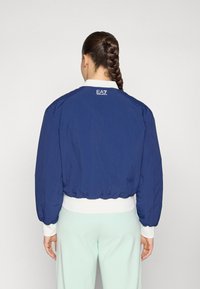 Navy bomber jacket with a ribbed white collar and cuffs, elastic hem, and logo detail on the back. Lightweight, smooth fabric texture.