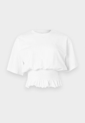 Λευκό κοντό τοπ με κοντά μανίκια και φαρδιά μανίκια, με σφηκοφωλιά στη μέση και βολάν στο τελείωμα, εκτίθεται σε ανοιχτό φόντο.