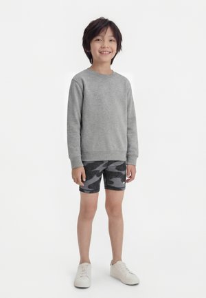 Usmievajúci sa chlapec s tmavými vlasmi, oblečený v sivej mikine s dlhým rukávom, čiernych športových šortkách s maskáčovým vzorom a bielych teniskách na bielom pozadí.