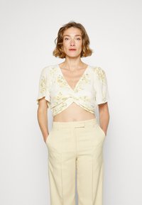 Top corto branco com estampado floral e mangas curtas e volumosas, apresentando uma frente torcida. Combinado com calças beiges de cintura alta, enfatizando um caimento descontraído.